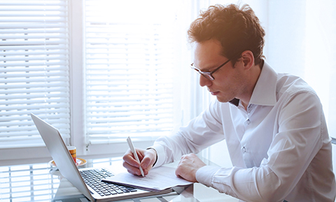Brighly lit room with a seated man wearing glasses, taking hand-written notes in front of an open laptop screen