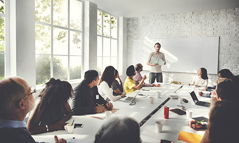 Office meeting space with many people seated around a long table, looking towards a man standing in front of a white-board, presenting information