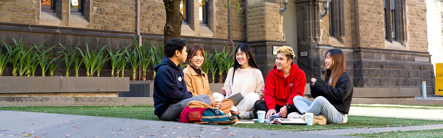 A group of students sitting together on grass in front of a historic campus building, talking and studying with notebooks and coffee cups.