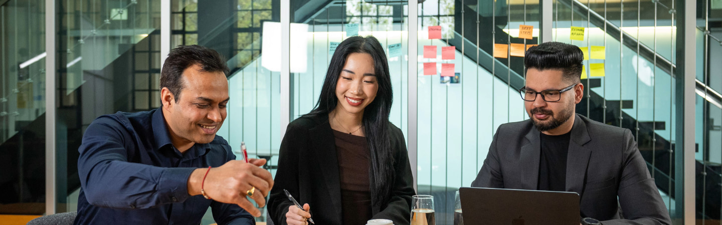 Three postgraduate students inside a modern office. The man on the left is writing, while the other two students watch.