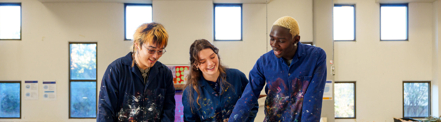 Three students wearing navy paint covered smocks in an art studio looking down and smiling as they lean in and one seems to gesture at an unseen artwork on a table in front of them.
