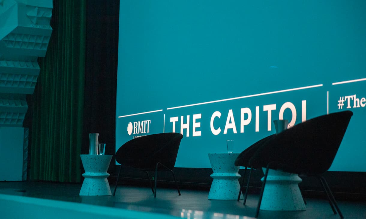 A close up photo of an empty stage with two empty chairs and three white stools. The stage is washed in a dark blue light the words The Capitol are projected on the screen behind the chairs.