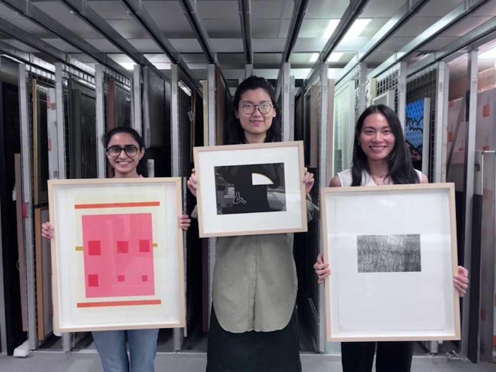 three smiling students holding up framed artworks from the artothek collection