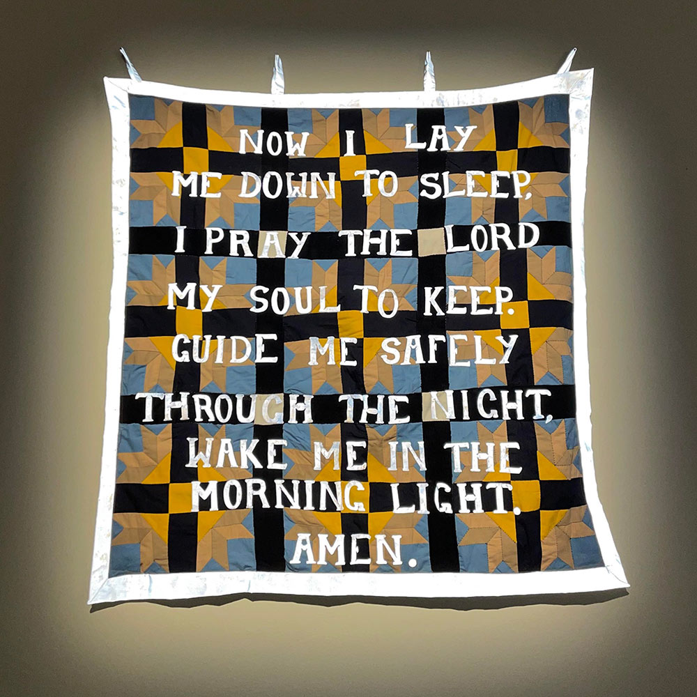 A quilted fabric that has blue, black and yellow checks – the yellow part almost makes the pattern of a sun. Over the top of the check is text stitched onto the fabric in white which reads ‘now I lay me down to sleep, I pray the lord my soul to keep. Guide me safely through the night, wake me in the morning light. Amen’.