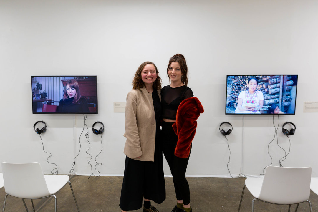 Two female students standing and smiling in front of two screens in gallery