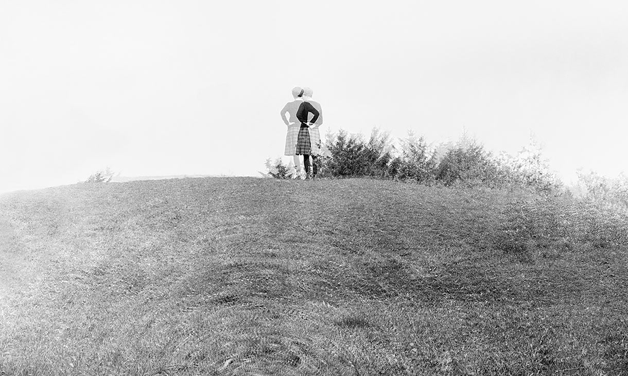 Black and white image in which the subjects look displaced – there is a person wearing a jumper and kilt standing on top of a hill with arms on their hips, looking away from the camera.