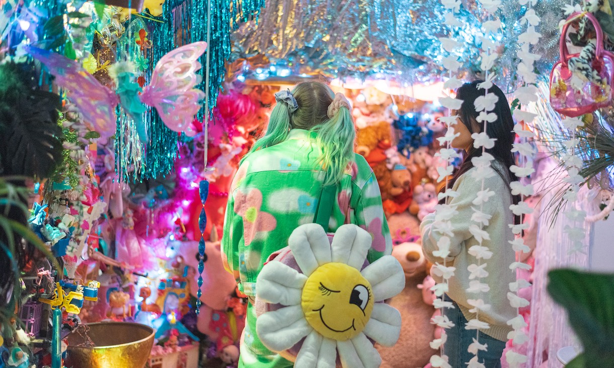 A person wearing a green, pink and white jumpsuit and a flower bag – the face on the centre of the flower is winking. There are lots of brightly-coloured lights and toys surrounding the person. 