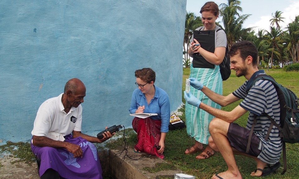 On the remote Fijian island of Naviti, RMIT staff (A/Prof. Matt Currell) and students (Liv and Alex) train community elder (Tai Samsi) how to sample drinking water from the blue communal rainwater tank behind (Source: Nick Brown)