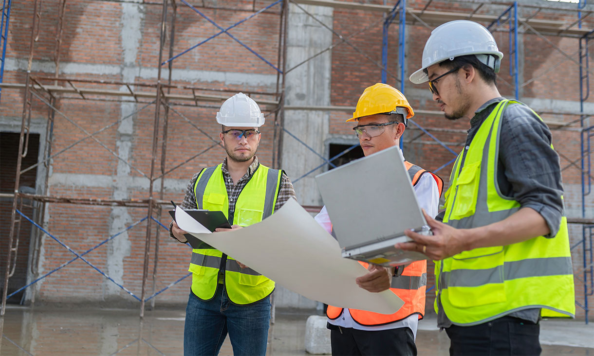 Three construction engineers wearing hard hats reviewing plans and data on a building site.