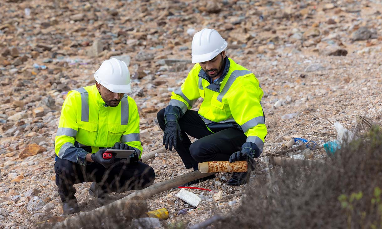 Two engineers in high-visibility jackets examining soil samples on rocky ground.