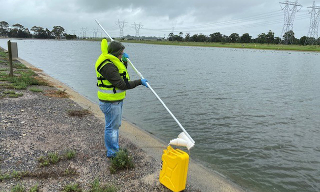 A person wearing a bright yellow safety vest and blue gloves, standing at the edge of a large body of water, using a long pole to collect a water sample. High-voltage power lines are visible in the background.