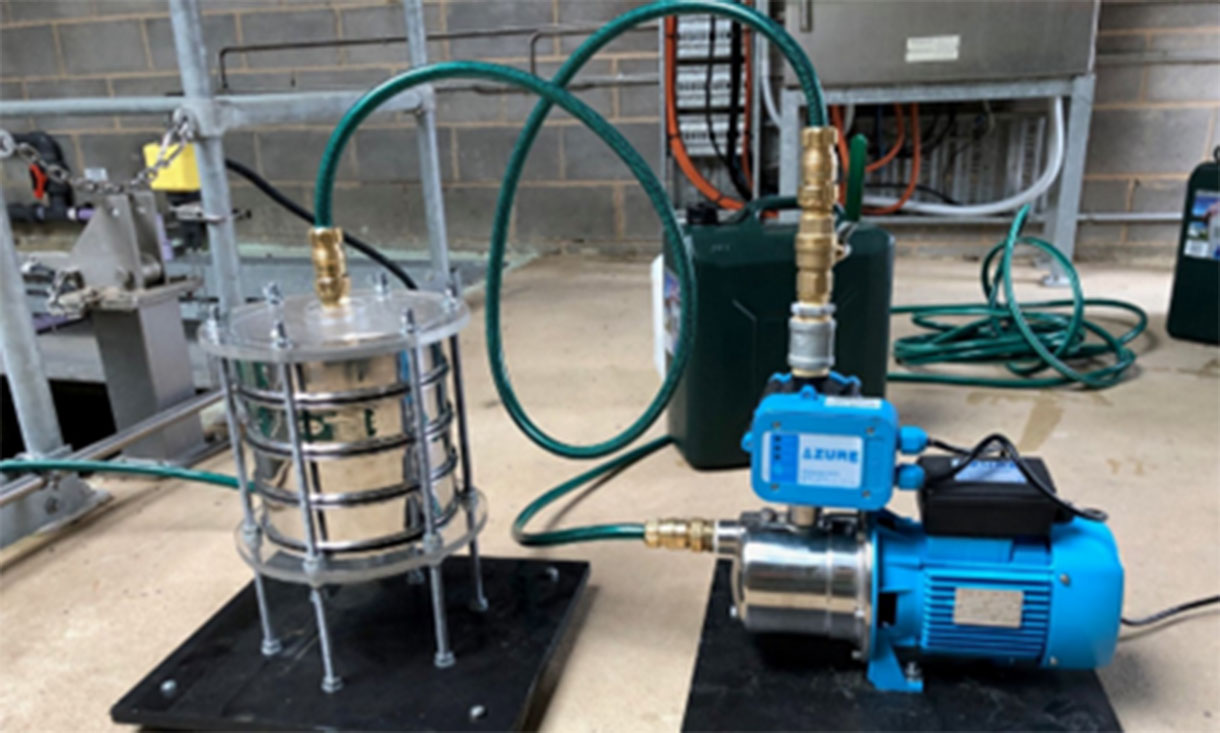 Close-up of industrial equipment used in water treatment. The setup includes a metallic cylindrical vessel connected to a pump with blue and green hoses, positioned on a cement floor.