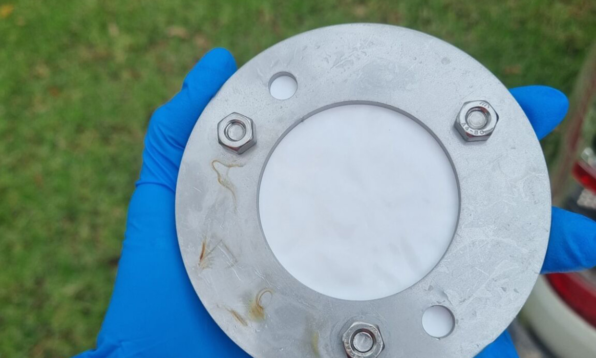 A close-up view of a circular metal component held by a person wearing blue gloves. The component has bolts and a smooth surface, with blurred greenery in the background.