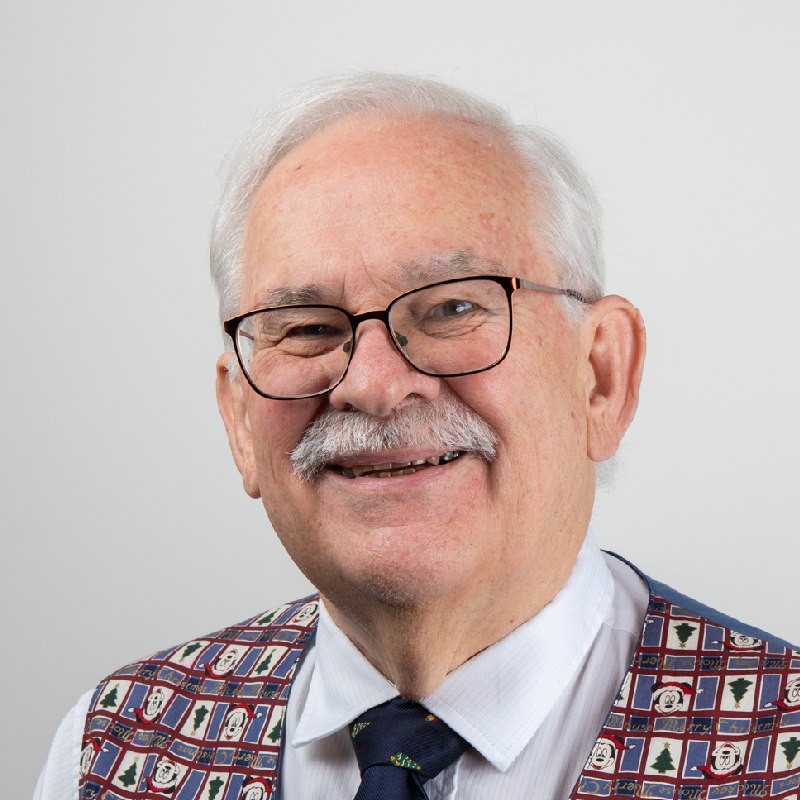 Stephen is smiling towards the camera against a solid white background. Stephen is wearing a white shirt and dark necktie underneath a patterned waistcoat.