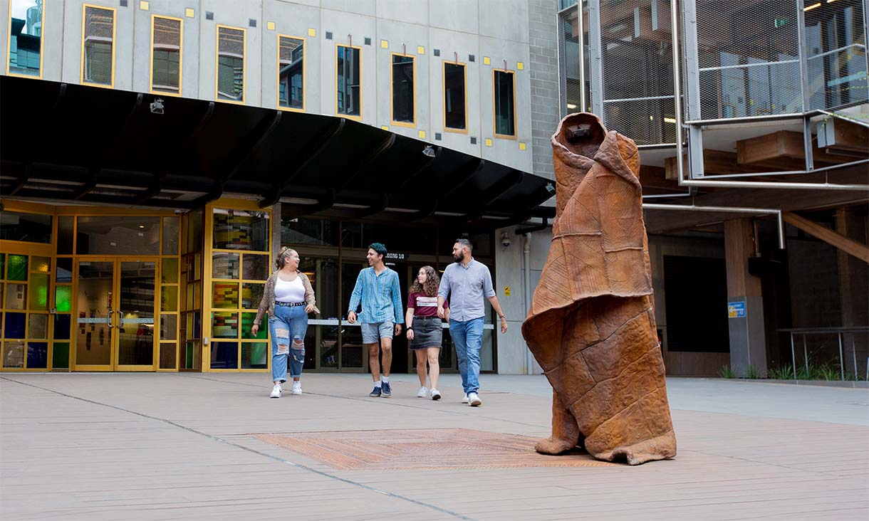 Group of students walking on the RMIT campus near a large, abstract brown sculpture in front of a modern building with yellow-framed glass doors and windows.