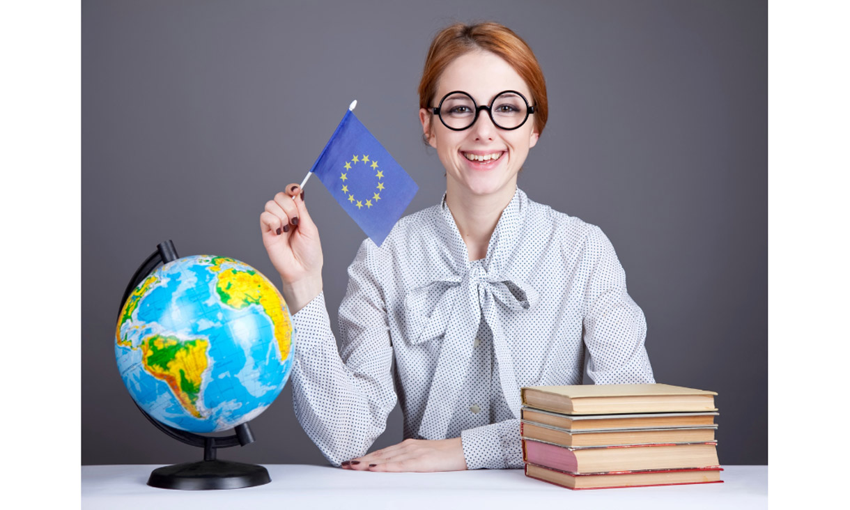 Photo of a person sitting behind a desk. The left of the desk has a globe that is facing America and Africa, and the right of the desk has five books stacked on top of each other with the spine not being visible. The person is wearing a white long sleeved shirt with black dots, black glasses and is holding an EU flag in their right hand. Their left hand is resting on the desk. They are looking at the camera straight on and smiling.