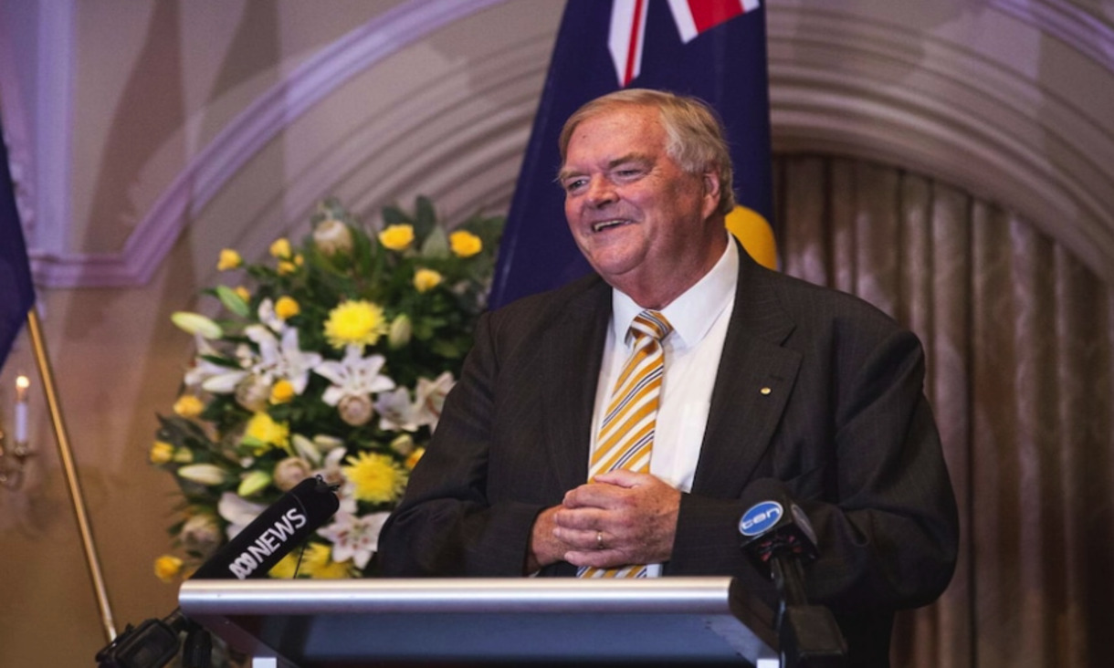 Former labor leader Kim Beazley standing at lecturn with Australian flag and white and yellow flowers behind him