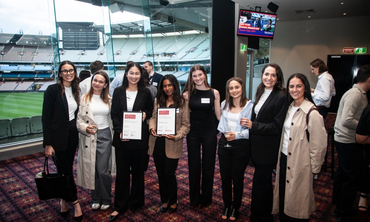 A group of eight smiling women stand in a glass box above the MCG stadium, two holding award certificates.