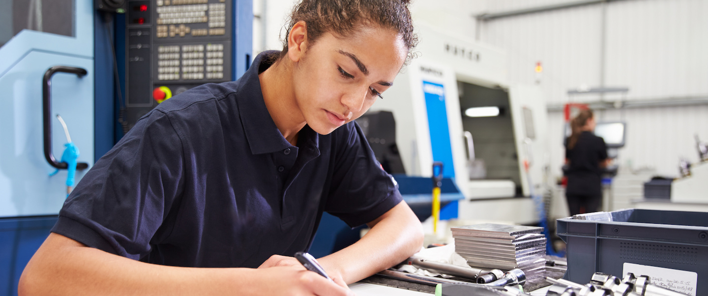 a young female engineer writing notes with a CNC machine in the background and industrial equipment at the desk