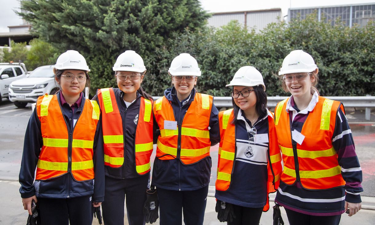 group of smiling female high school students wearing high vis vests and hard hats posing for photo on excursion at asphalt plant