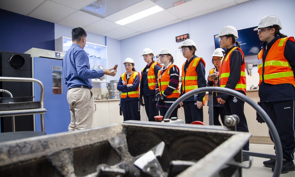 group of female high school students wearing high vis vests and hard hats listening to presenter in a lab on excursion at asphalt plant