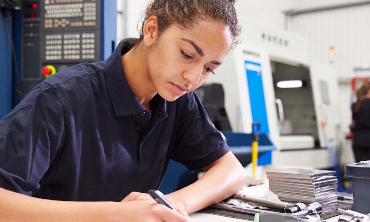 a young female engineer writing notes with a CNC machine in the background and industrial equipment at the desk
