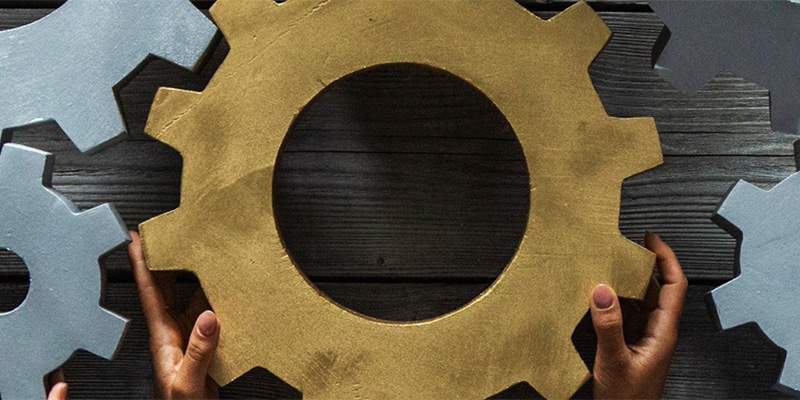Hands holding a large gold cog, surrounded by silver cogs on a wooden surface.