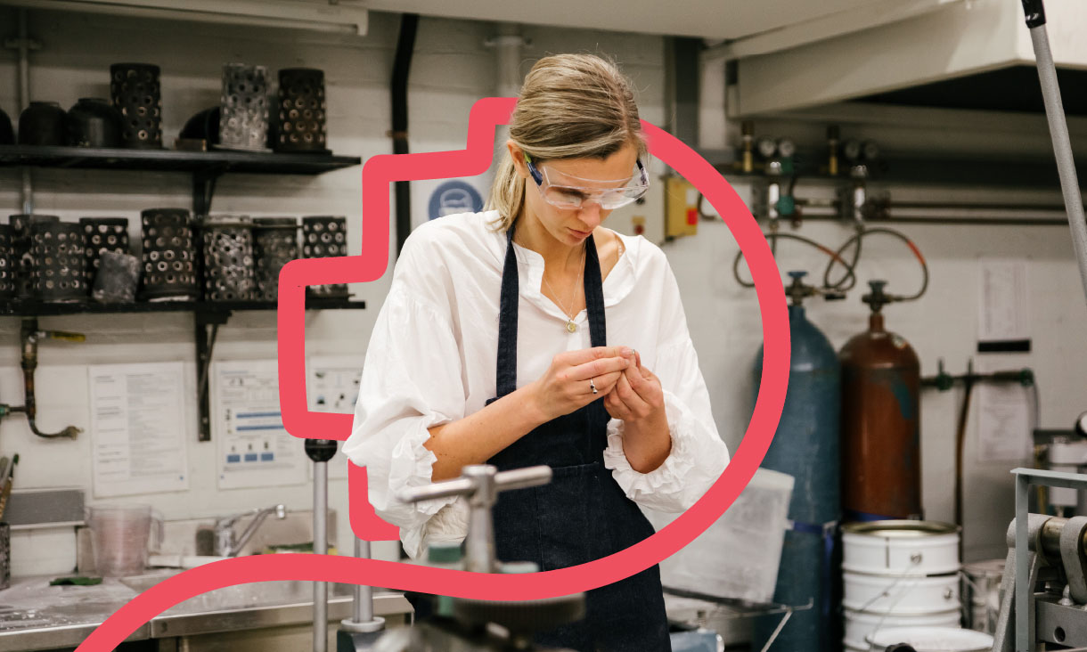 Person in a white shirt and apron working in an industrial workshop surrounded by equipment and shelves.