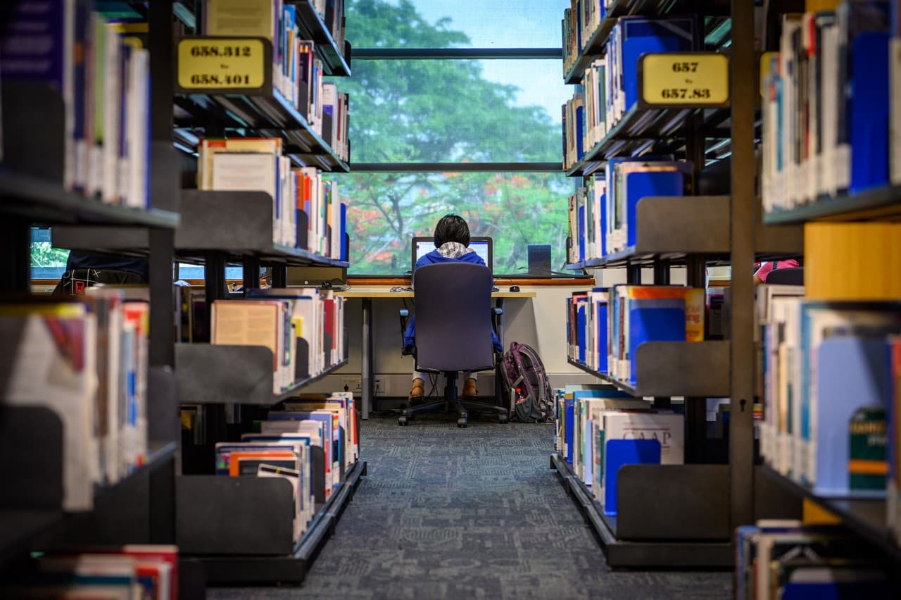 A student sitting at a desk in a library with bookshelves surrounding.
