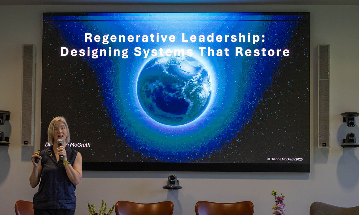 A presenter stands in front of a large screen that displays an image of Earth. The slide reads 'Regenerative Leadership: Designing Systems That Restore.' 