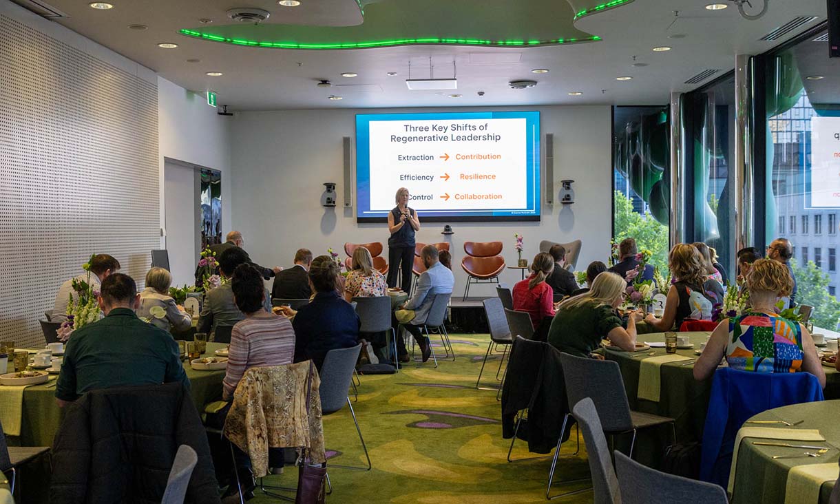 A speaker stands at the front of a conference room addressing an audience seated at round tables. Attendees are listening to a presentation. The room is well-lit, decorated with green accents and floral centrepieces.