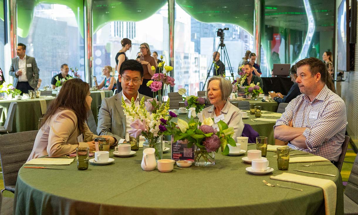 Small groups of people sit around round tables in a contemporary meeting space, engaging in conversation. The tables are set with cups, glasses and floral arrangements.
