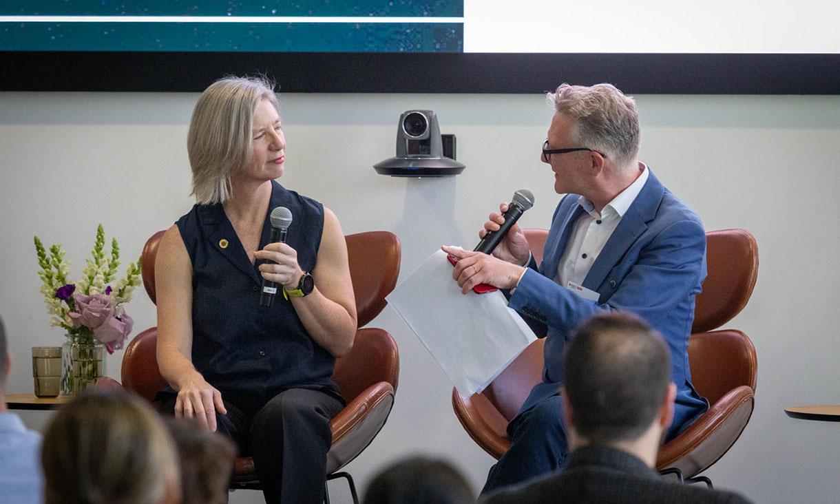 Two people seated on stage having a discussion with microphones during an event, with an audience in the foreground and a flower arrangement on a table nearby.