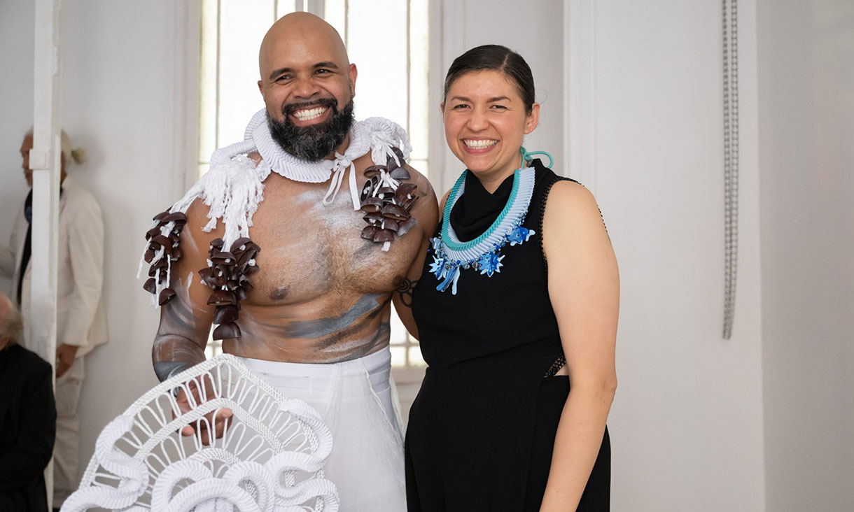 Two people standing together in a bright room, wearing bold, sculptural garments featuring intricate beadwork and textured designs, one person holding a decorative white piece.