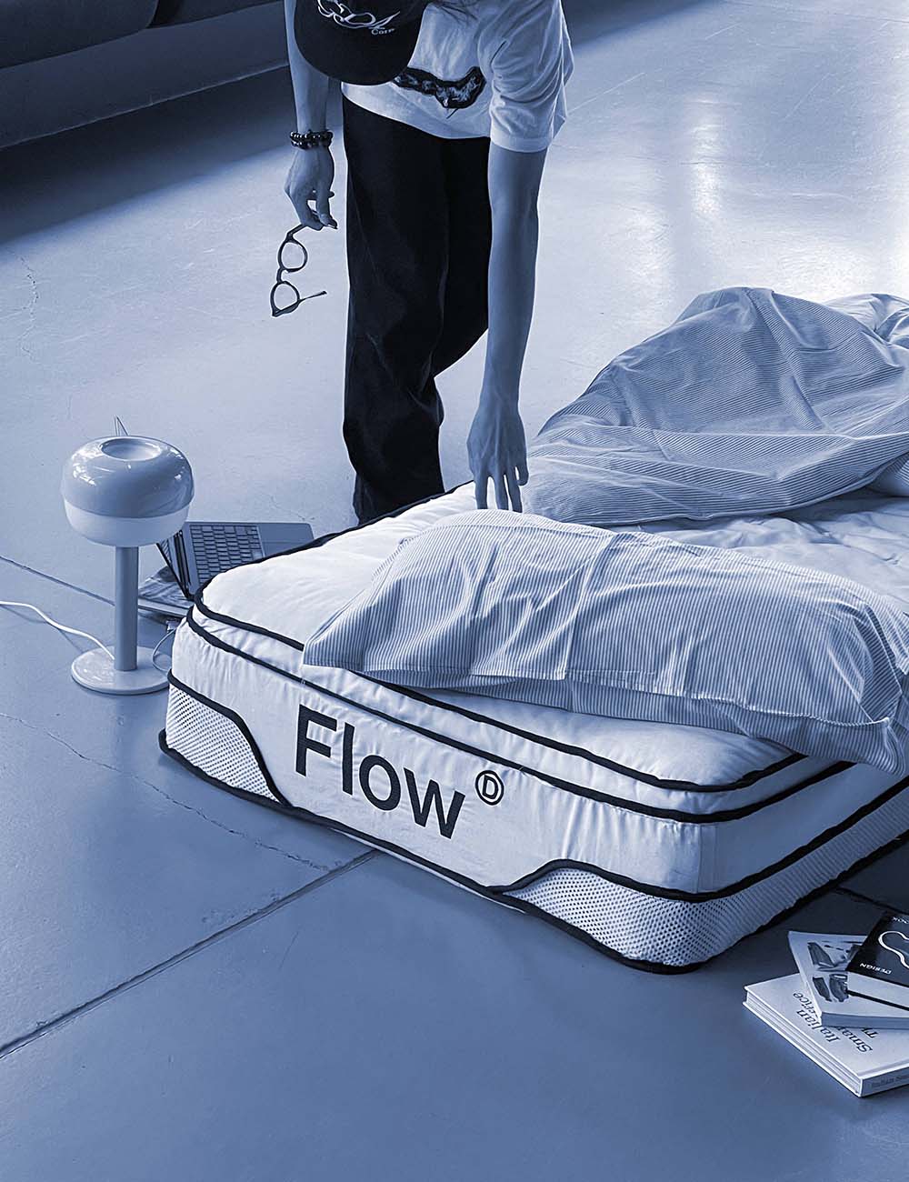 Person making a bed with striped sheets, next to a mattress labeled 'Flow', with a modern lamp and open laptop on the floor.