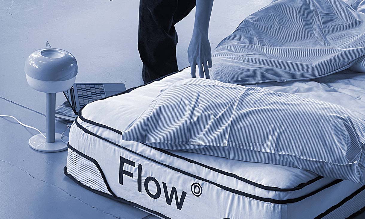Person making a bed with striped sheets, next to a mattress labeled 'Flow', with a modern lamp and open laptop on the floor.