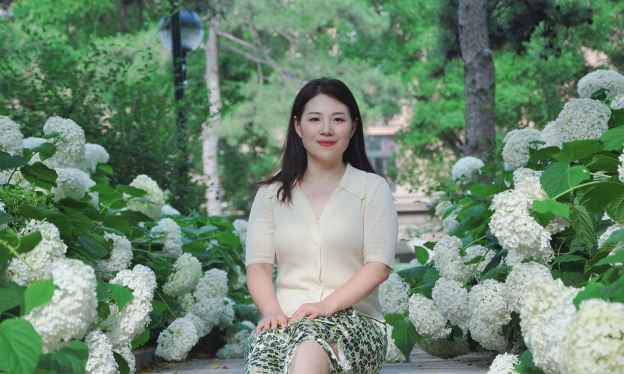 RMIT alumni Jing Wei, sitting in a garden of large white flowers, wearing a white cardigan and green patterened skirt.
