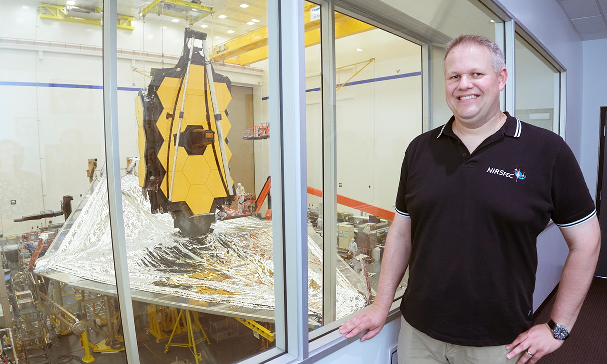 RMIT alumnus Marc stands smiling with a piece of aerospace equipment visible through windows beside him.