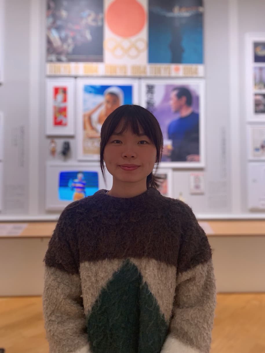 Minxuan photographed wearing a brown sweater standing in a gallery space