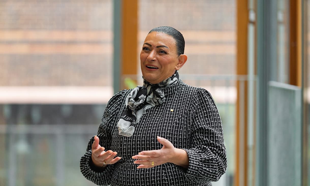 Rosanna Baini is pictured speaking and gesturing in a well lit glass room, wearing a black and white patterned outfit.