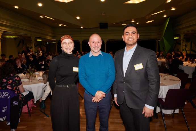 Bethany is on the left wearing a black turtleneck top, headscarf and gold-rimmed glasses. Bradley is in the middle wearing an aqua jumper and Adil is on the right wearing a grey suit jacket and blue shirt. They are all smiling and you can see people standing and sitting at tables in the background.