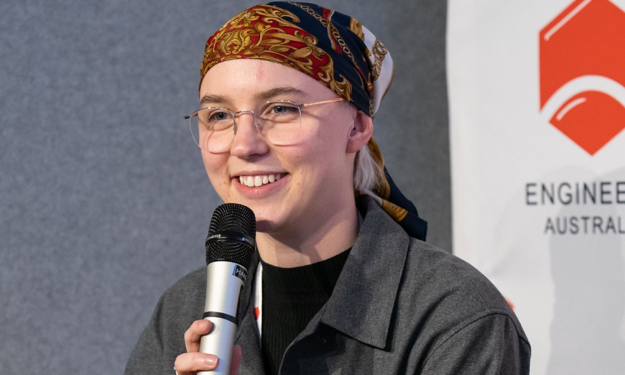 Close-up of Bethany holding a microphone and smiling. She has short white hair and is wearing a colourful headscarf. She is also wearing gold framed glasses and a grey jacket.