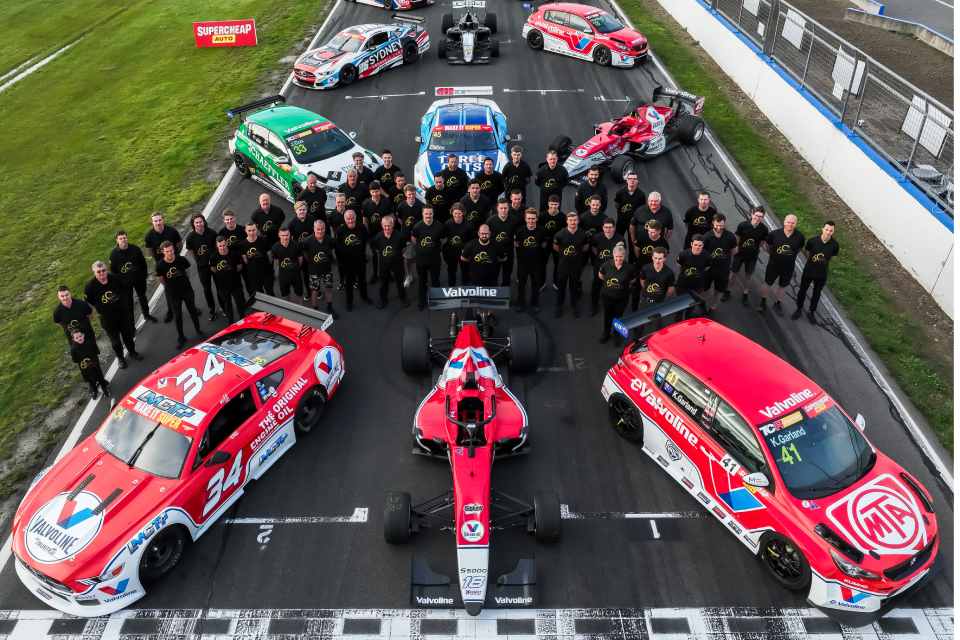 An aerial shot of the Gary Rogers Motorsport team standing on a racetrack. They are all wearing black shirts and pants and are surrounded by twelve race cars.