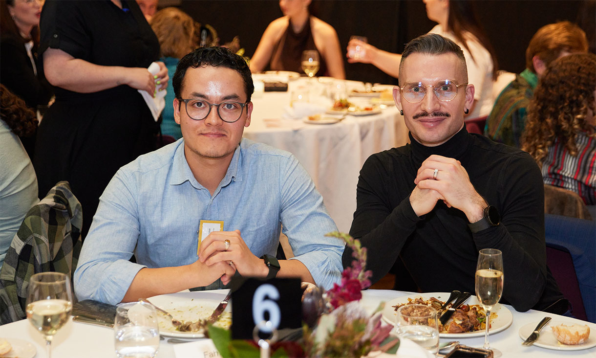 Fahim has short dark hair that curls at the front, and he is wearing a pale blue shirt and round glasses. Sitting to his right, Robbie Van Dijk has short brown hair, a thin moustache and gold rimmed glasses. He is wearing a black turtleneck jumper. They are sitting at the dinner table and there are other guests in the background.
