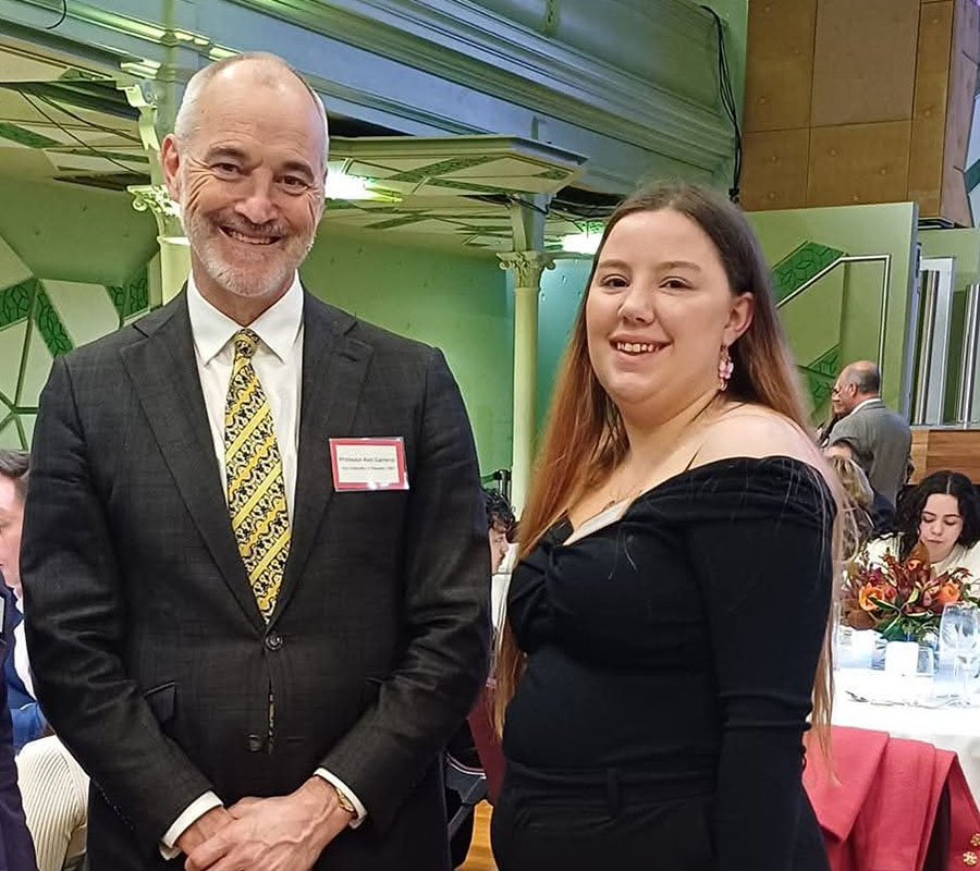 Isabelle is wearing a black dress and is standing to the right of Professor Cameron who is wearing a grey suit, white shirt and yellow patterned tie.