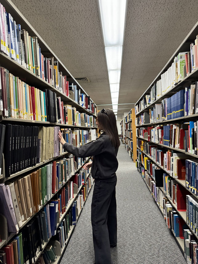 Expanding knowledge: Louise King delving into fashion and communications resources at the Fashion Institute of Technology library.    Alt text: Louise is standing in a long row of bookshelves in a library.