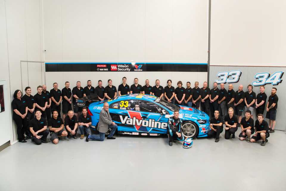 The Gary Rogers team of thirty six people are all wearing black shirts with a blue logo. They are standing around a blue race car with Valvoline written on it in white.