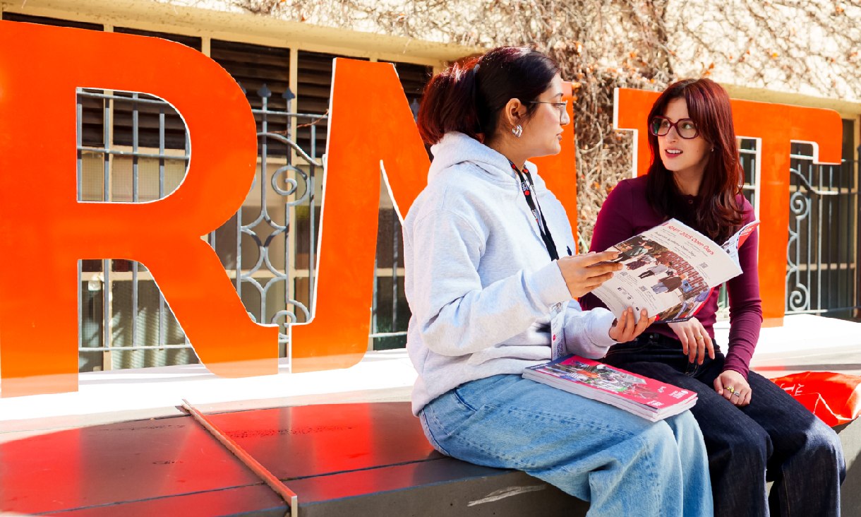Two students sitting and talking in front of large, bright orange RMIT University letters, holding course guides and study materials outdoors on campus.