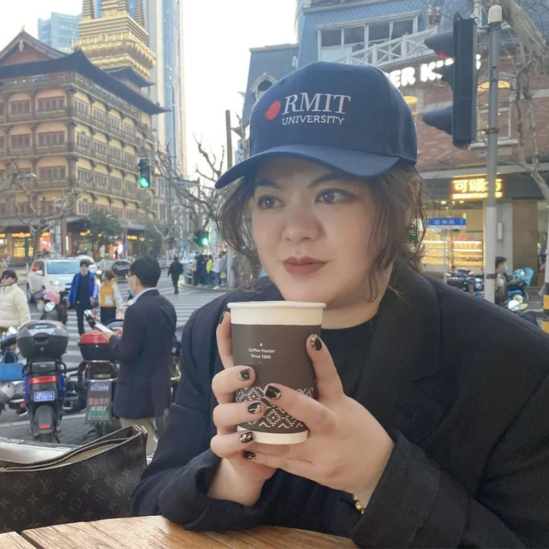 Cindy pictured sitting at a cafe drinking a coffee wearing an RMIT hat, with a bustling street behind her.