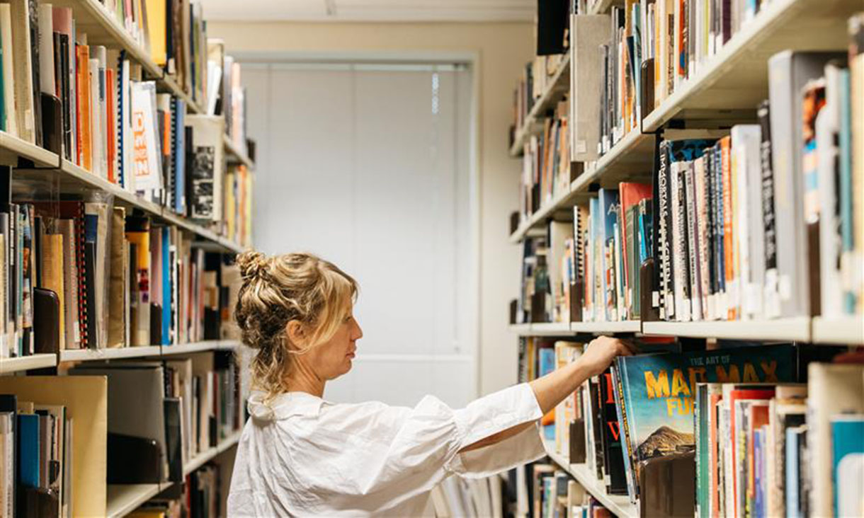 A person with light hair in a bun is standing in a library, reaching for a book on a shelf lined with various colourful books. The environment is bright, with shelves full of books extending into the background.
